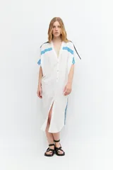 Túnica blanca de lino con corte oversized, escote en V, cierre con botones y mangas cortas. Presenta bordados geométricos azules en hombros y laterales.