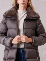 Campera puffer larga de acabado satinado en tono beige. Presenta un diseño capitoneado horizontal, cierre frontal bidireccional y un amplio cuello envolvente revestido en piel sintética. Incluye bolsillos laterales funcionales con cierre y logo metálico en la manga.