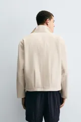 Cazadora de tejido color crema, con cuello alto, cierre frontal de cremallera y bolsillos de vivo en la cadera. Presenta un calce relajado (relaxed fit).