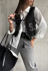 Chaleco de gamuza negra con bordados decorativos en color blanco y cierre frontal mediante cordones con borlas. Presenta un diseño corto y estilo boho.