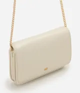 Cartera bandolera pequeña marca Satinato, color beige, confeccionada en poliuretano con diseño estructurado, solapa frontal y detalle de logo metálico dorado. Cuenta con correa larga de cadena metálica dorada.