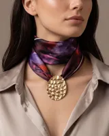 Pañuelo de seda con estampado multicolor en tonos violeta y burdeos, acompañado por un gran dije metálico dorado de textura martillada.