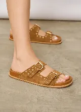 Sandalia chata de gamuza color marrón, con dos tiras cruzadas en el empeine, hebillas de metal dorado y detalles de ojales metálicos a lo largo de las tiras.
