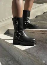 Botas de caña media en cuero negro, con suela track dentada y taco cuadrado. Presentan un diseño con correas ajustables, argollas metálicas y detalles de tachas plateadas.