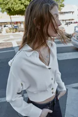 Camisa corta de cuello solapa y manga larga, con trabillas decorativas en los hombros. Presenta pliegues delanteros, elástico en la espalda y cierre frontal con botones.
