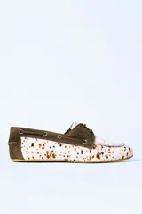 Mocasín tipo náutico de cuero con pelo, con diseño estampado de manchas en tonos marrones y blancos. Presenta detalles en gamuza marrón en el talón y la zona de los cordones, con ojalillos metálicos laterales.