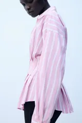 Camisa de popelina con diseño a rayas verticales rosadas y blancas, cuello solapa con escote en V, manga larga y detalle de pliegues marcados en la cintura. Cierre frontal con botones ocultos.