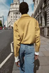 Chaqueta bomber de efecto ante en color tostado, con cuello solapa, manga larga y bolsillos laterales. Presenta un diseño con bajo acabado en pliegues que aporta volumen y cierre frontal con cremallera y botones a presión.