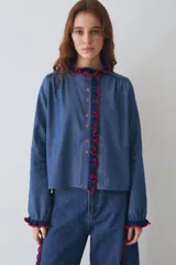 Camisa de algodón estilo denim con corte regular, cuello redondo y cierre frontal con botones. Presenta detalles de volados tejidos a mano en crochet en color azul marino con bordes rojos en el cuello y la cartera frontal.