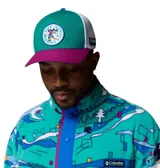 Gorra Columbia x Disney tipo trucker, con panel frontal de color verde, visera fucsia, paneles traseros de malla blanca y parche redondo con gráfico de Mickey Mouse practicando esquí. Incluye etiqueta lateral con el logo de Columbia y Disney.