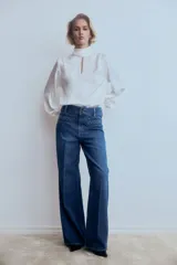 Pantalón de jean estilo wide leg con tiro alto, confeccionado en denim de algodón rígido. Presenta bolsillos delanteros con detalle de ribete trenzado, pliegues frontales y traseros, y cierre con doble botón.