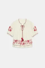 Blusa color crudo de manga corta con bordados en rojo a contraste en el cuello, mangas y ruedo. Cuenta con escote redondo con abertura y cordones con borlas colgantes.