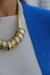 Collar de diseño artesanal compuesto por múltiples tiras de cuero en color crudo y piezas cilíndricas de metal dorado con textura martillada.