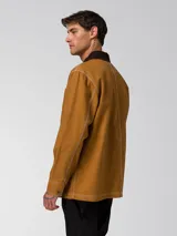 Chaqueta tipo chore de lona color marrón, con cuello camisero en contraste color marrón oscuro, cierre frontal con botones, cuatro bolsillos de parche frontales y costuras a la vista en color blanco.
