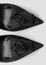 Zapato de tacón destalonado color negro de tejido satinado, con punta fina, cierre de hebilla ajustable en la tira del talón, tacón de aguja y detalle de flor decorativa en el empeine.