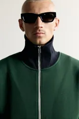 Chaqueta regular fit color verde oscuro con detalles en negro en el cuello subido, los puños, el dobladillo inferior y los ribetes de los bolsillos. Cuenta con cierre frontal de cremallera metálica y bolsillos con cierre en la cadera.