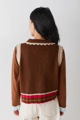 Chaleco de lana color marrón con cuello estilo bebé, cierre frontal con botones grandes y detalle de puntilla en cuello, sisas y bolsillo frontal. Presenta una franja decorativa en color rojo en el ruedo.