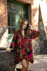 Vestido corto de lino con estampado floral en tonos fucsia, verde y negro. Presenta un diseño asimétrico, más corto en el frente y más largo en la parte posterior, con escote en V y mangas amplias.