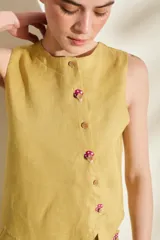 Top de lino blanco sin mangas, con cuello redondo y cierre de botones de madera dispuestos en una cartera asimétrica. Cuenta con delicados detalles bordados en forma de flor alrededor de los ojales.
