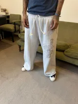 Pantalón de jogging gris de corte baggy, confeccionado en algodón. Presenta cintura elástica con cordón largo ajustable, botones en la parte inferior para ajustar la botamanga y un diseño artístico que combina estampa en plastisol con bordado en relieve sobre el muslo izquierdo.