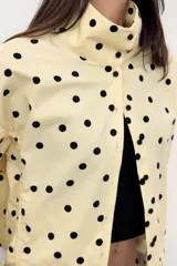 Chaqueta de cuello subido y manga larga con estampado de lunares negros sobre base beige. Cuenta con bolsillos de vivo en los laterales, detalles de frunces ajustables con cordones en el delantero y cierre frontal con botones.