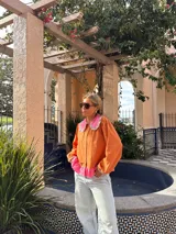 Camisa de manga larga color naranja con cuello grande en contraste color rosa y detalle de volados en el borde. Presenta un diseño con botones frontales y un detalle de tela rosa en la parte inferior.