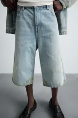 Bermudas de denim de algodón, color celeste claro con efecto lavado y manchas marrones/beige en las perneras. Tienen un corte ancho (baggy fit) y terminación de bajo irregular.
