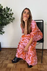 Vestido largo con estampado de hojas tropicales en tonos naranja y rosado. Presenta escote en V, mangas cortas abullonadas con ajuste de lazo y falda con volados en el ruedo.