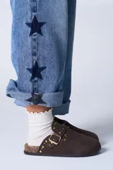 Zueco de gamuza color marrón chocolate, con detalle de tachas doradas y hebilla metálica ajustable en el empeine. Presenta una base de corcho y suela de goma antideslizante.