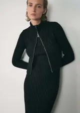 Vestido midi de punto canalé color negro, con diseño recto y manga larga. Presenta un cuello perkins con cierre de cremallera frontal que recorre la parte superior, creando un efecto de chaqueta superpuesta.