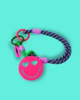 Llavero para bolso con cuerda trenzada en colores violeta y verde, decorado con un dije circular de acrílico rosa con diseño de carita feliz con ojos en forma de corazón. Incluye argollas metálicas de colores y una etiqueta pequeña de silicona.