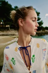 Vestido largo estilo túnica de color crudo con estampado multicolor de motivos marinos y mediterráneos. Presenta cuello con volante, cierre frontal con botones, manga larga y cordón ajustable en el cuello con abalorios.