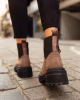 Borcego de gamuza color chocolate con paneles laterales elásticos negros y suela track dentada. Presenta sistema de cordones con ganchos metálicos y tirador en la parte trasera.