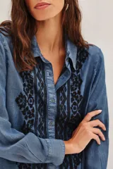 Camisa de denim azul con bordados negros en el frente, de manga larga, cuello clásico y cierre frontal con botones.