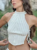 Top de tejido de punto color beige claro, con cuello alto y diseño acanalado vertical. Presenta aberturas laterales que se ajustan con lazos anudados.