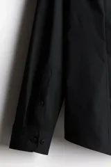 Camisa de vestir negra con cuello inglés, tapeta clásica y mangas largas con puños ajustables. Presenta un corte entallado que se ajusta al cuerpo y bajo levemente redondeado.