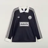 Camiseta Adidas de manga larga en colaboración con la banda Oasis, de color negro con cuello polo, puños y detalles en blanco marfil. Presenta las tres tiras características en las mangas, el logo trébol de Adidas, un escudo circular conmemorativo de la gira 'Live '25' y el estampado con la inscripción 'oasis' en el centro del pecho.