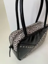 Cartera rígida negra con detalles en rojo y tachas plateadas. Tiene herrajes y cierre metálico plateado marca YKK, asa fina para llevar al hombro o en la mano y correa larga extra regulable. El interior es forrado, tiene un bolsillo con cierre enmarcado en cuero, y un segundo bolsillo plaqué.