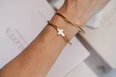 Pulsera de esferas doradas con dije central en forma de cruz de color blanco nacarado.
