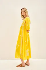 Vestido largo hasta los tobillos de color amarillo, con bordados florales blancos a contratono. Presenta mangas tres cuartos, escote redondo con cartera de botones frontales y un diseño holgado.