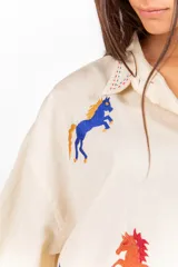 Camisa de corte clásico y calce amplio, color marfil, con cuello camisero, mangas largas y cierre frontal con botones. Presenta bordados coloridos de caballos en contraste distribuidos en la prenda.