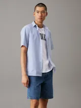 Camisa de manga corta Calvin Klein confeccionada en popelín de algodón elástico con diseño de rayas verticales en blanco y celeste. Presenta cuello en punta, cierre frontal de botones, bolsillo de parche en el pecho y bajo recto.