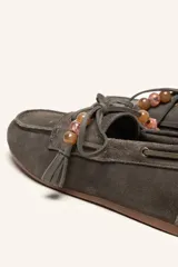 Mocasín estilo náutico de gamuza marrón, con cordones de cuero, borla decorativa y cuentas de colores en el empeine.