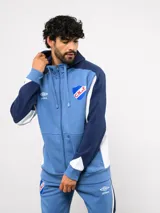 Campera deportiva Umbro del Club Nacional de Football, con cierre frontal, capucha ajustable con cordones y diseño en bloques de color azul, celeste y blanco. Presenta el logo de la marca y el escudo del club bordados en el pecho.