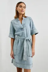 Vestido camisero corto de denim celeste con cuello en V, mangas cortas y cinturón de lazo. Presenta bolsillos laterales y pespuntes a contratono.