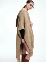 Ruana tejida de color beige con capucha integrada y terminaciones con flecos en el ruedo. Presenta un diseño envolvente y holgado, confeccionada en una mezcla de lana merino extrafina y viscosa.