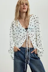 Top de crepe blanco con estampado de lunares negros, escote en V y cierre frontal con finas lazadas. Presenta mangas largas abullonadas con puños elásticos y bajo redondeado.