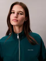 Campera tipo track Calvin Klein de tejido plano en color verde oscuro, de corte cropped y manga larga. Cuenta con cuello alto, cierre frontal de cremallera metálica, bolsillos laterales y elástico acanalado a rayas en los puños y el dobladillo. Presenta el logo de la marca bordado en el pecho.