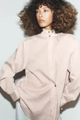Camisa de corte oversize con estampado de rayas verticales, cuello subido y cierre frontal con botones. Presenta mangas largas con puños y un diseño cruzado en la parte delantera.