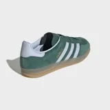 Championes Adidas Gazelle Indoor de gamuza verde oscuro, con las tres tiras características en color blanco y el nombre del modelo en letras doradas en el lateral. Poseen suela de goma translúcida y detalles en color celeste claro en el talón.